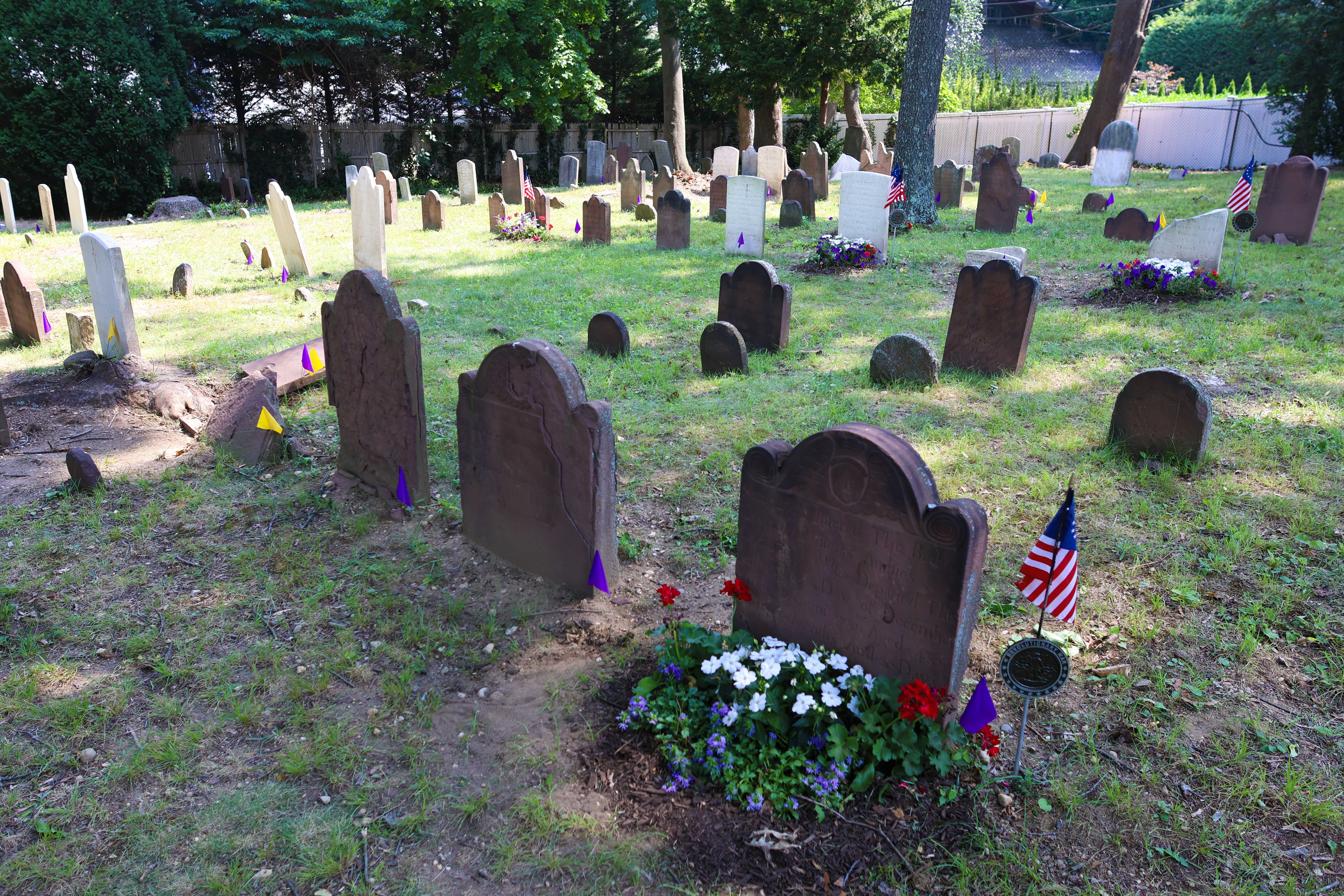 A wide view of the historic Monfort Cemetery in Port Washington, with restored Revolutionary War gravestones, American flags, and colorful memorial flower beds.