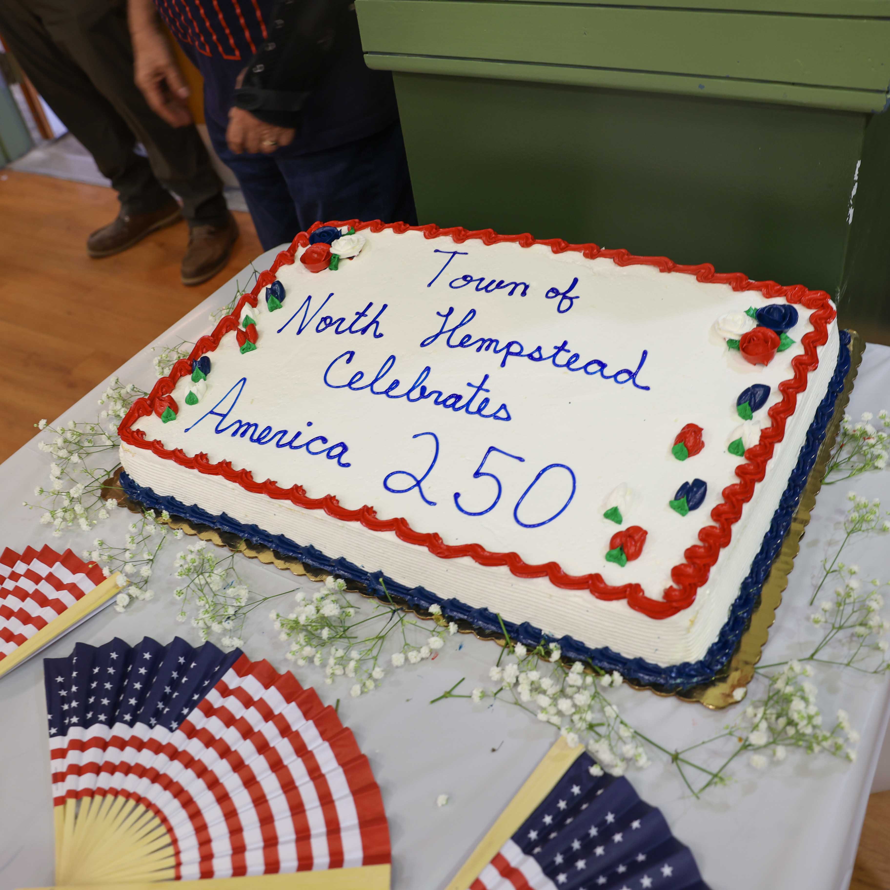 The America 250 celebration cake, decorated in red, white, and blue with the inscription 'Town of North Hempstead Celebrates America 250,' surrounded by American flags and patriotic fans.