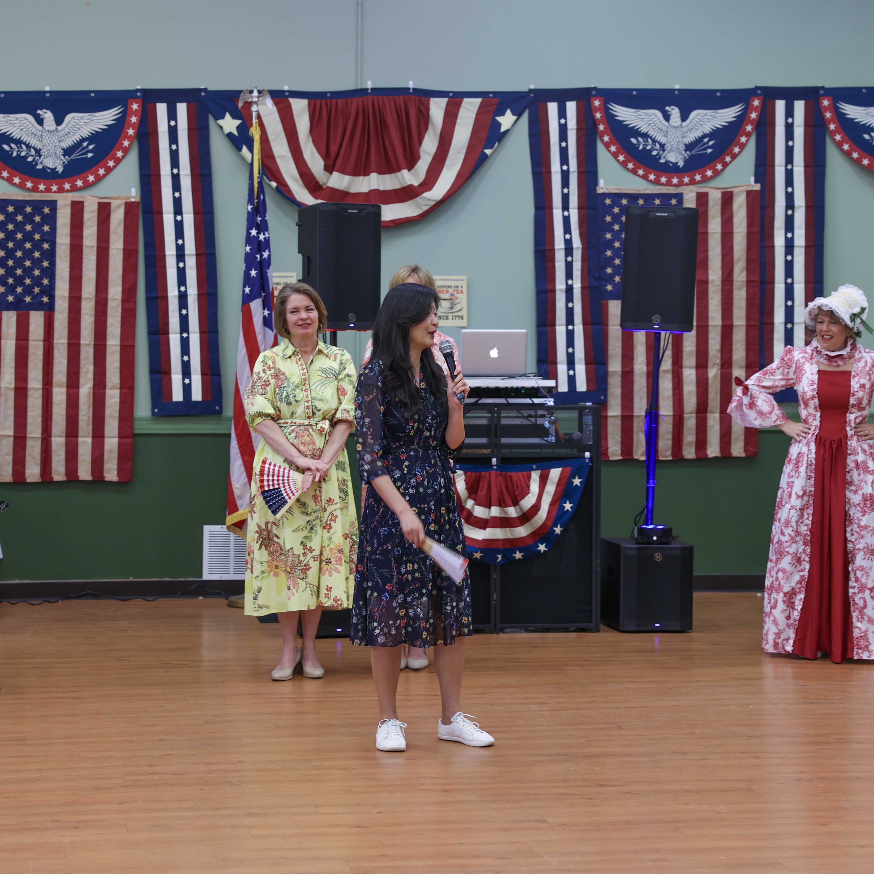 Supervisor Jennifer S. DeSena and Town Clerk Ragini Srivastava at the program, accompanied by a performer in colonial-era costume, with patriotic flags and bunting decorating the stage.