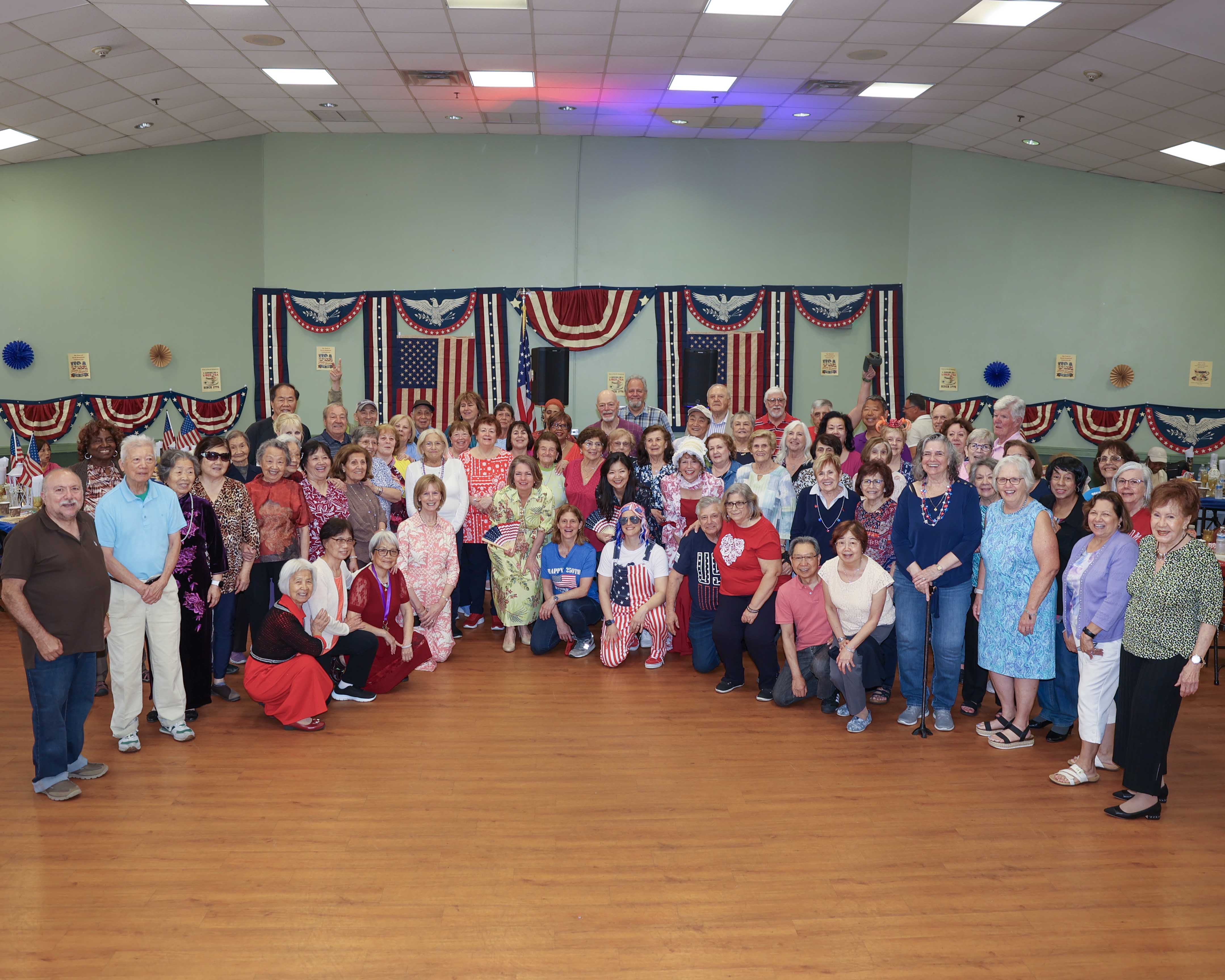 A large group photo of event attendees filling the dance hall, many dressed in red, white, and blue, with patriotic bunting and American flags across the back wall.