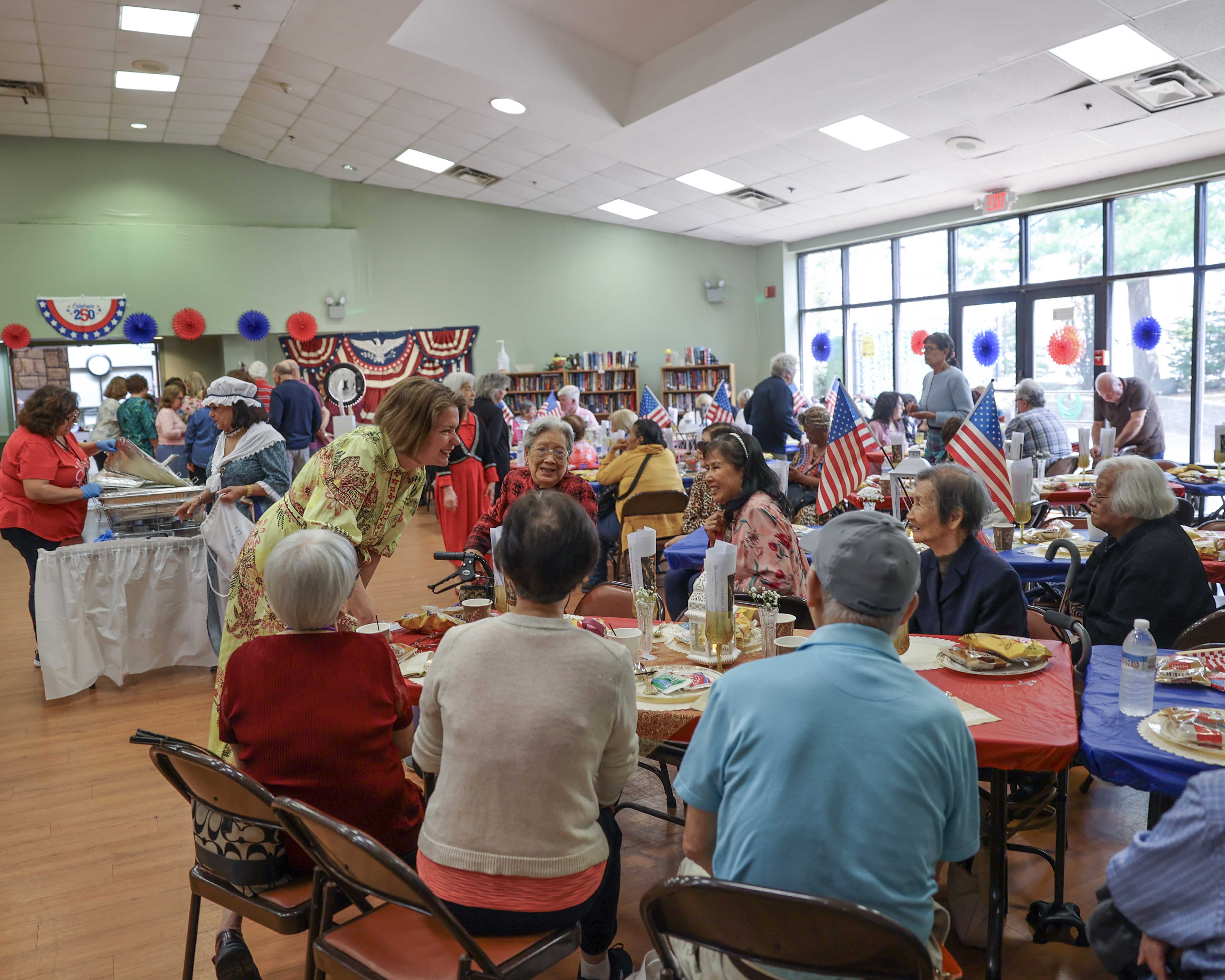 Supervisor Jennifer S. DeSena leans in to greet seniors seated at decorated lunch tables, with the full event hall visible behind her.
