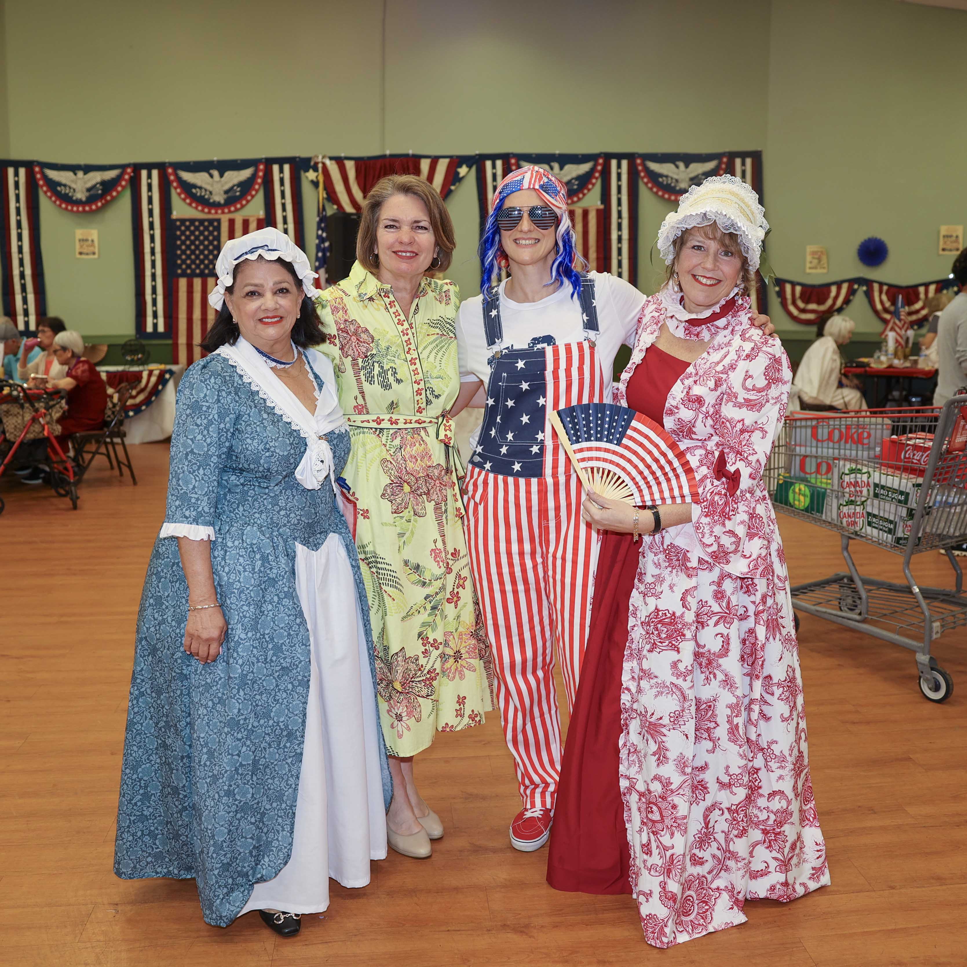 Four women in patriotic costumes — including two in colonial dress — pose together on the dance floor with the decorated event hall behind them.