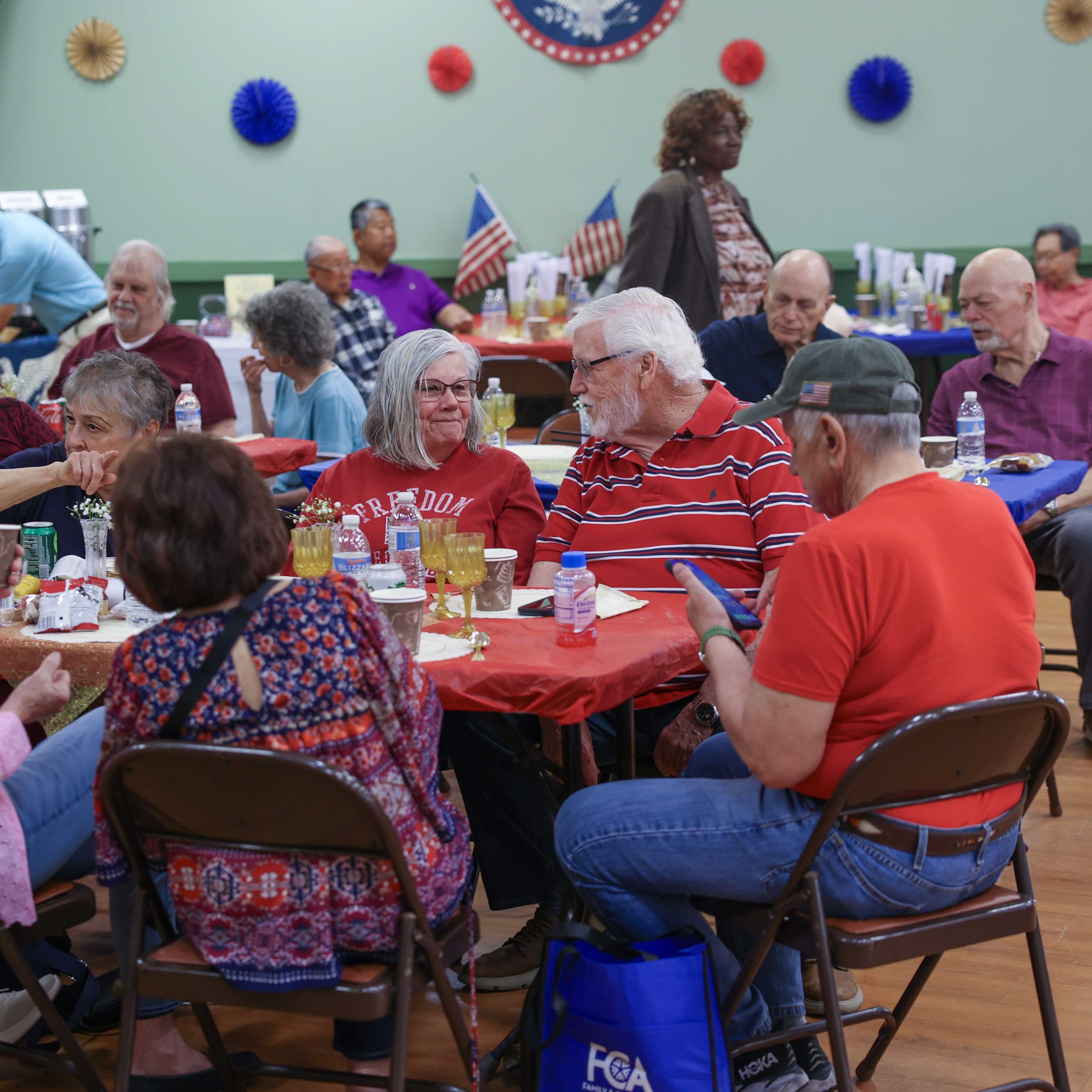Seniors seated at round tables enjoying lunch, decorated with red tablecloths, small American flags, and gold accents.