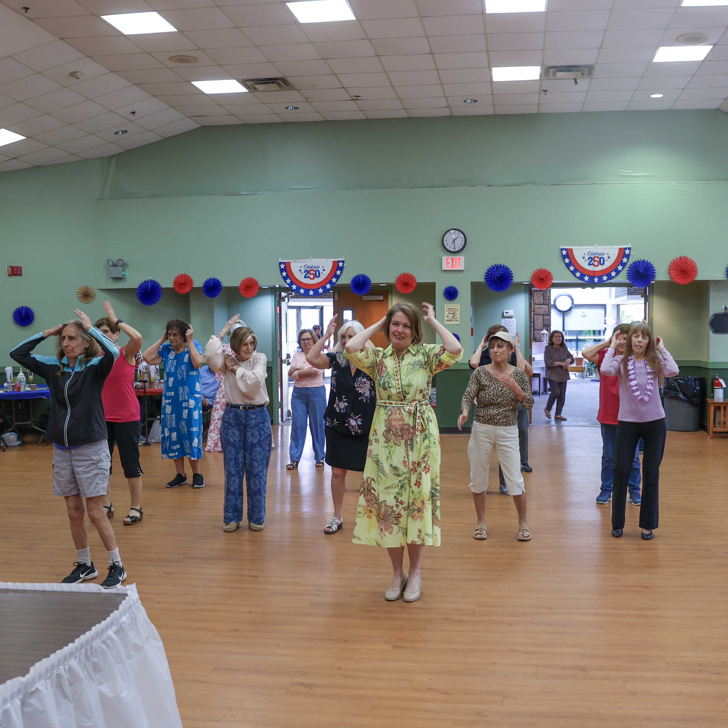 Supervisor Jennifer S. DeSena leads a line dance with seniors in the center of the hall, with 'Celebrate 250' banners visible above the doors.