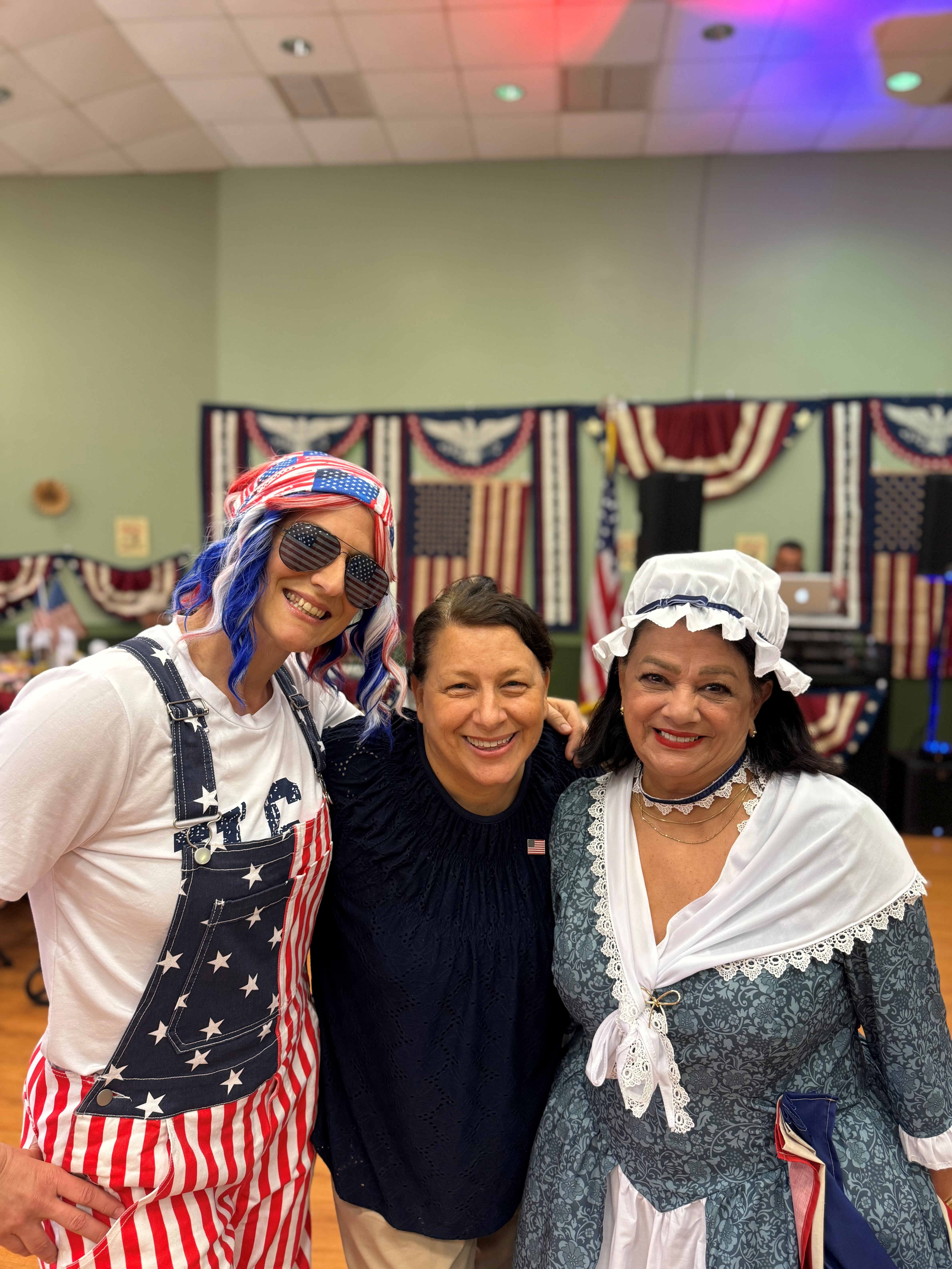 Three women in patriotic costumes smile for the camera, with the event's patriotic bunting and flags visible in the background.