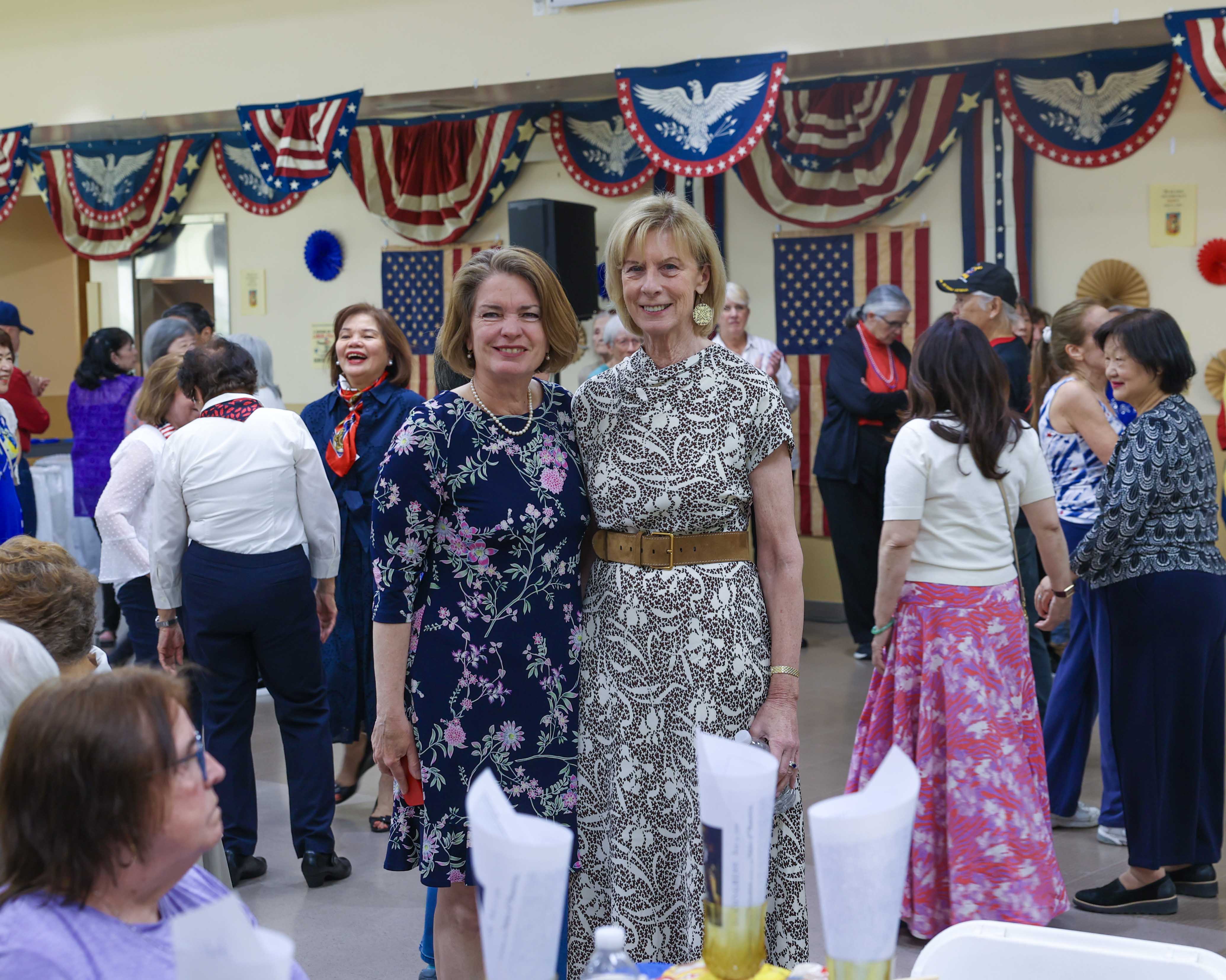 Supervisor Jennifer S. DeSena and Councilwoman Marianne Dalimonte pose on the dance floor as seniors dance behind them, with patriotic bunting and American flags decorating the hall.