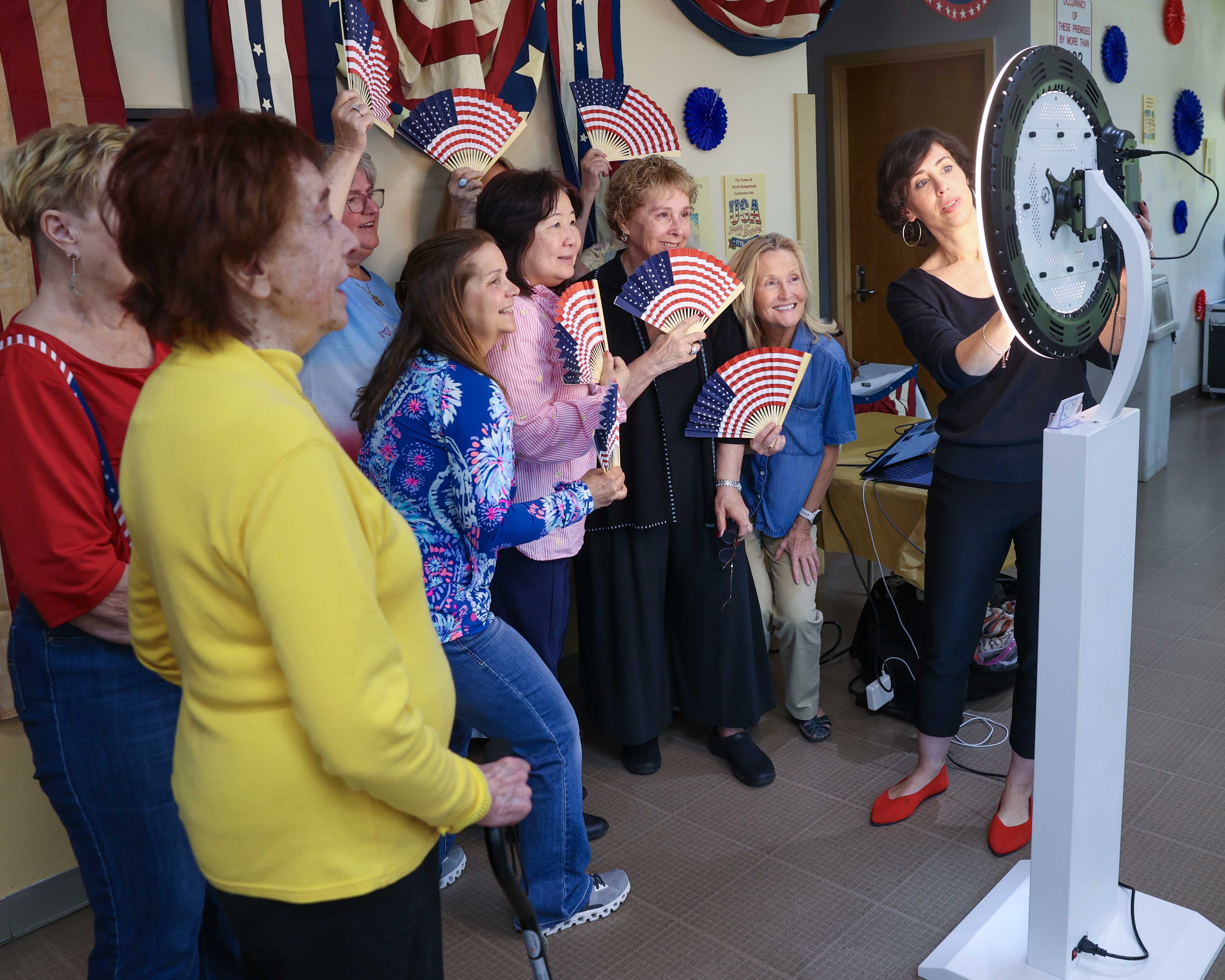 Seniors pose for a photo booth shot holding patriotic fans, guided by a staff member adjusting the ring light camera.