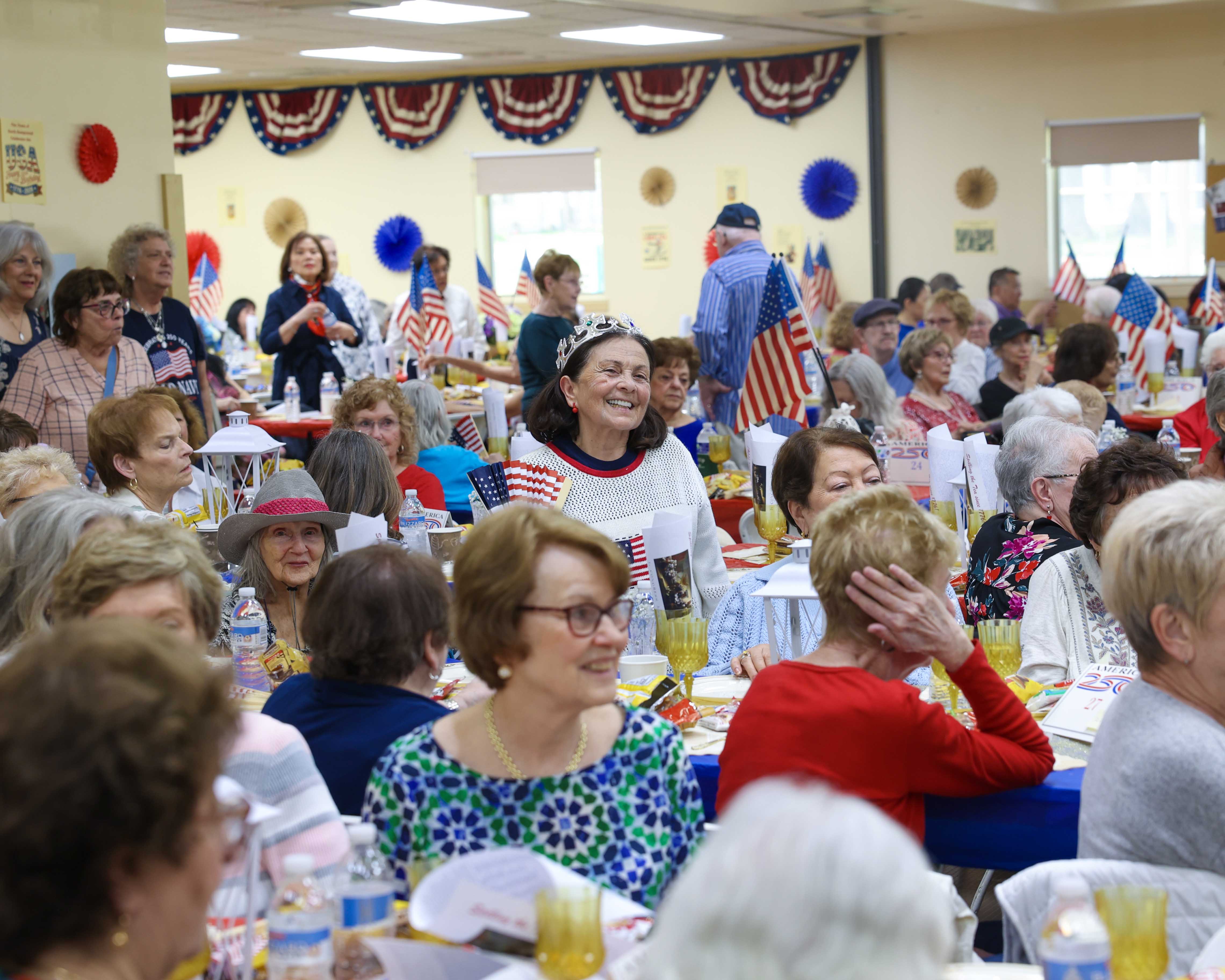 A packed room of seniors at the luncheon, tables decorated with American flags, gold accents, and America 250 centerpieces, with a woman in a tiara smiling in the center.