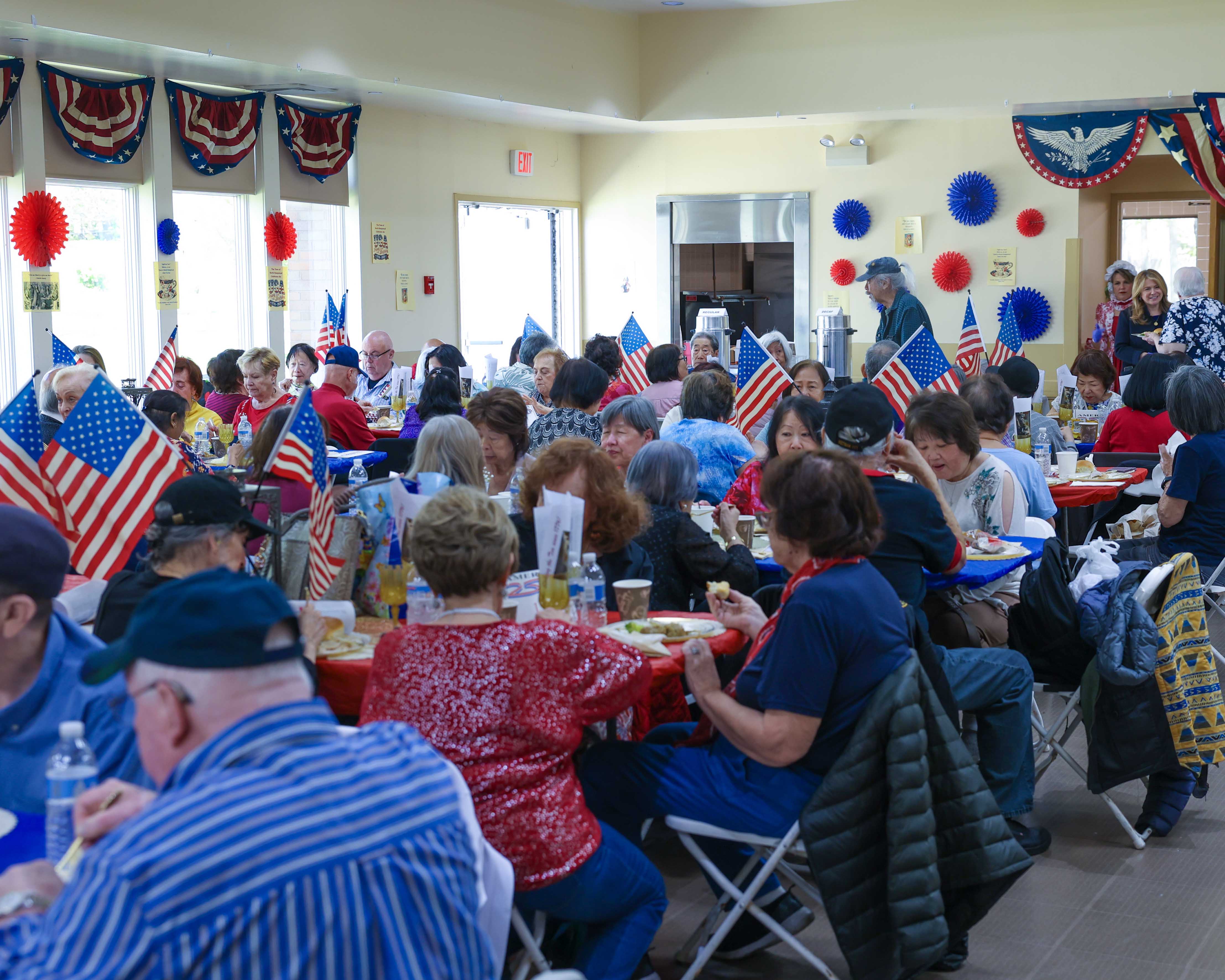 A full view of the luncheon hall with seniors seated at tables decorated with American flags, patriotic bunting lining the windows and walls.
