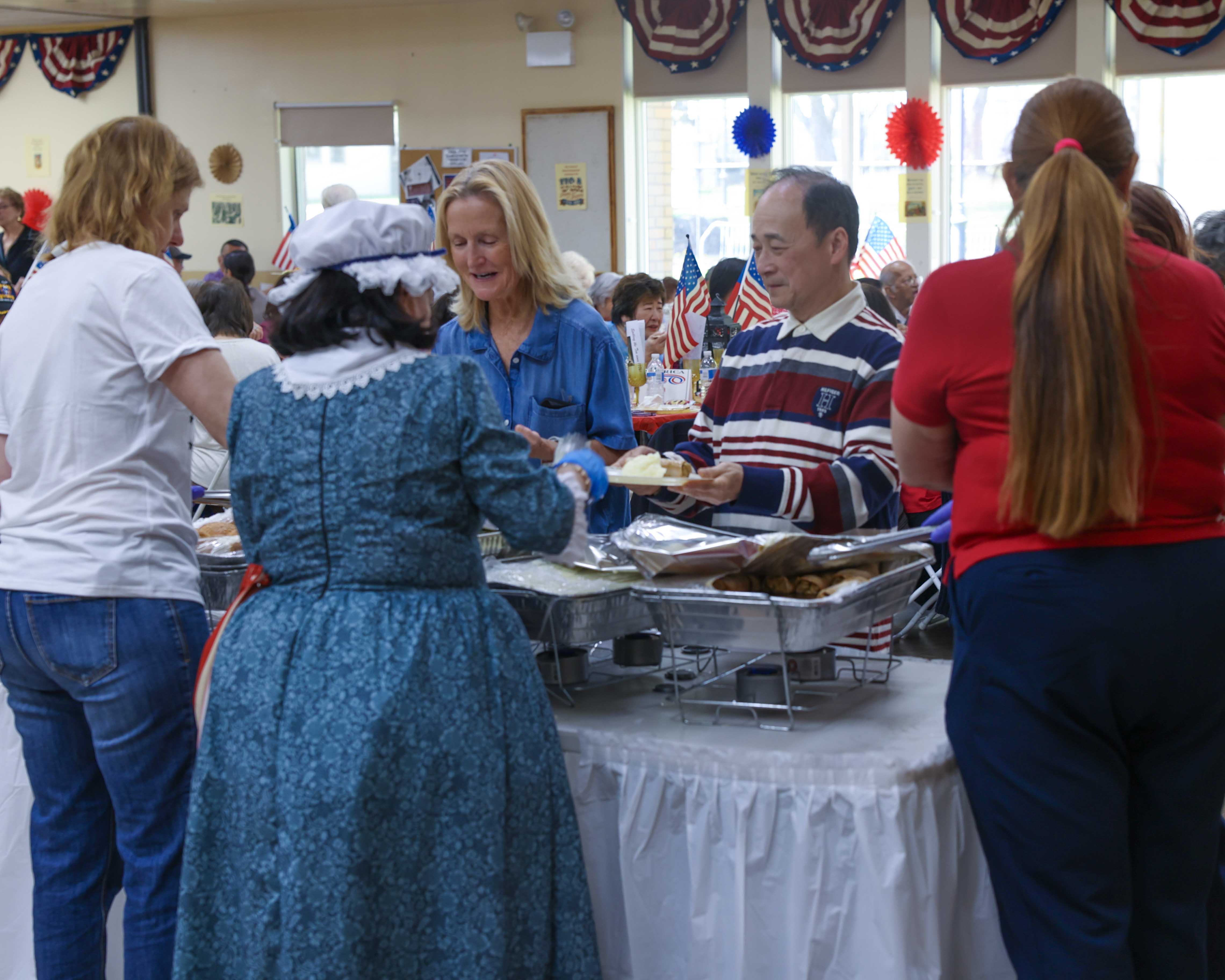 A volunteer in colonial costume serves lunch at the buffet table alongside other staff and attendees.