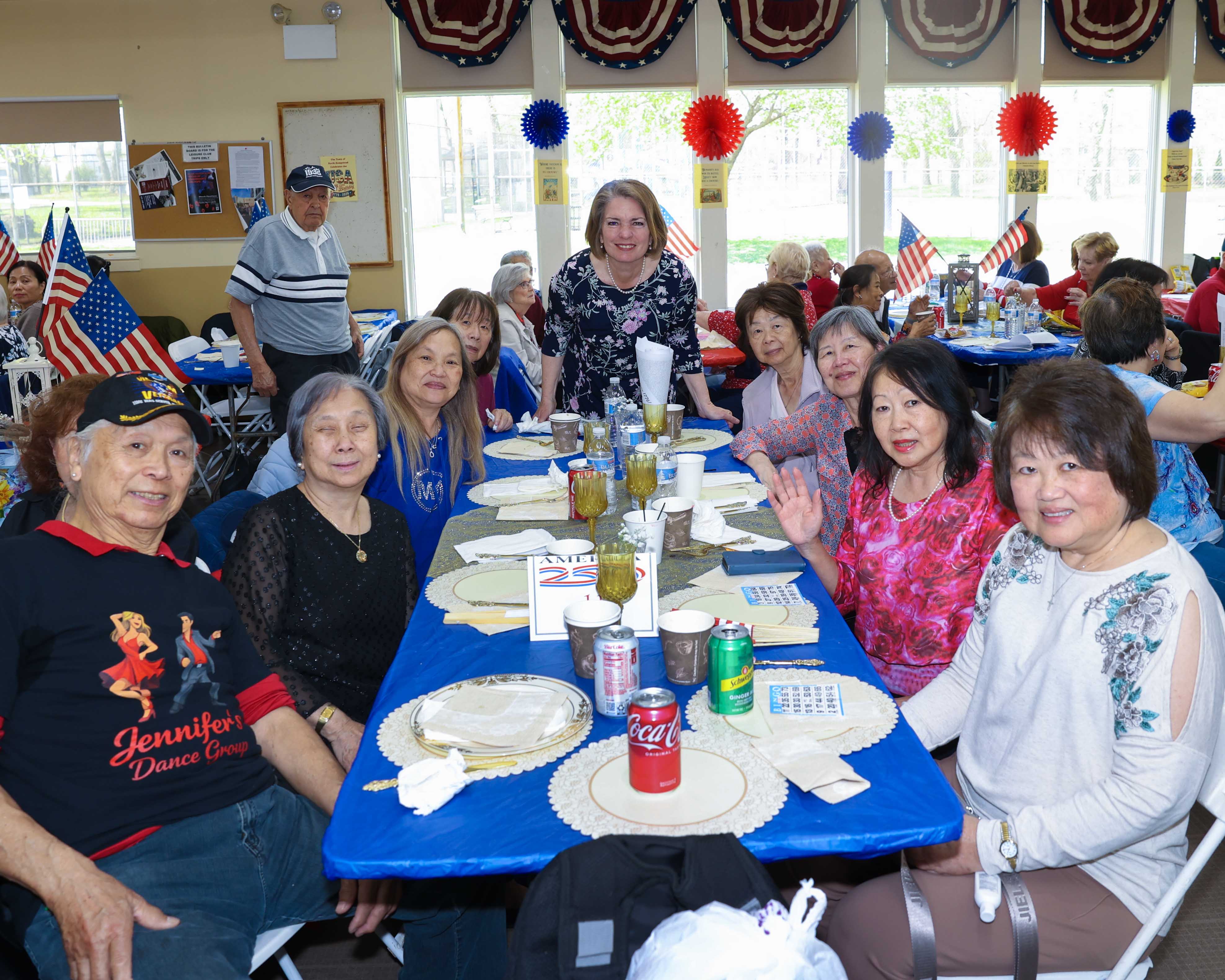Supervisor Jennifer S. DeSena stands with a table of seniors wearing Jennifer's Dance Group shirts, with an America 250 table centerpiece and American flags in the background.