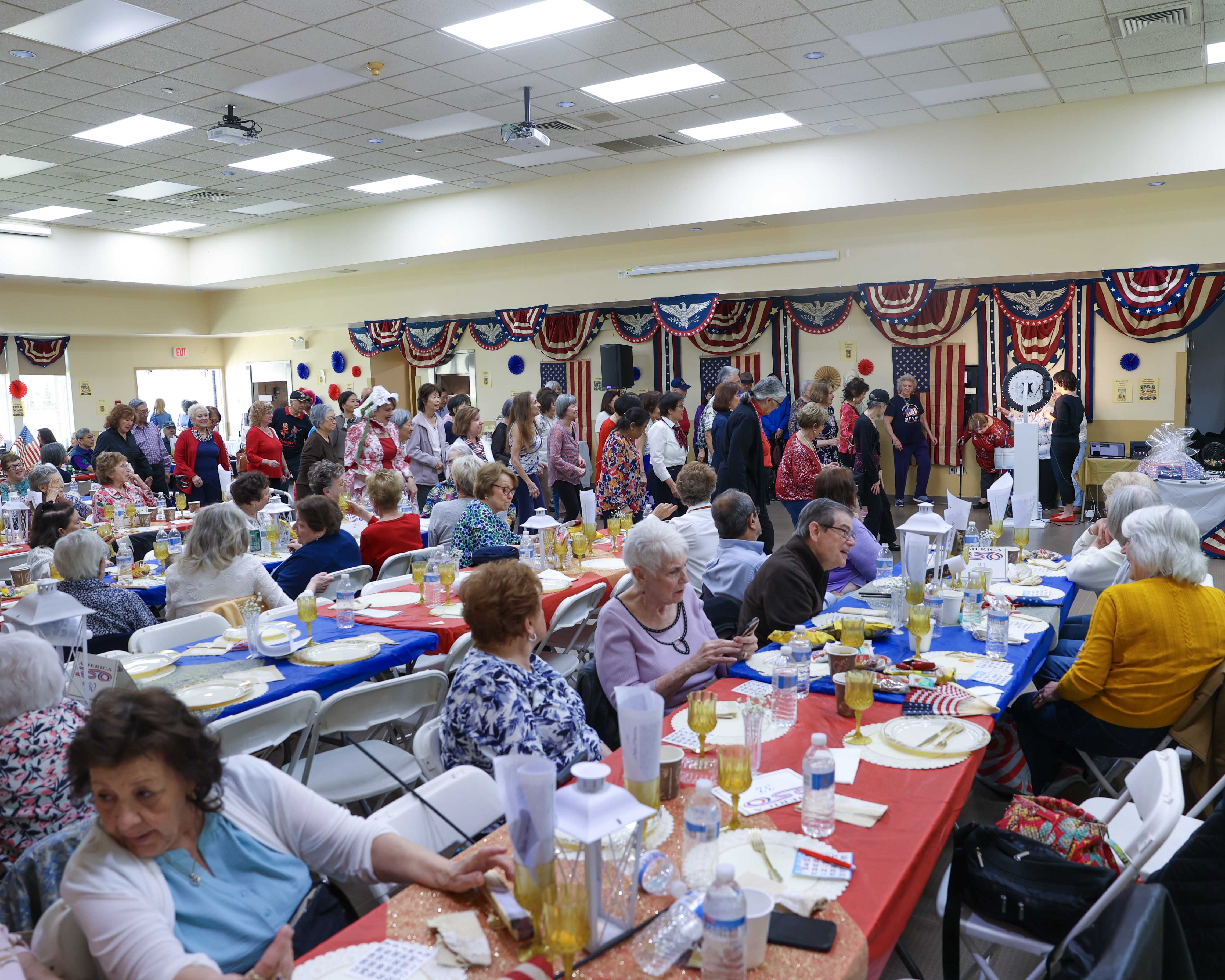 A wide view of the full event hall — seniors seated at long decorated tables while others line dance along the back wall beneath patriotic bunting.