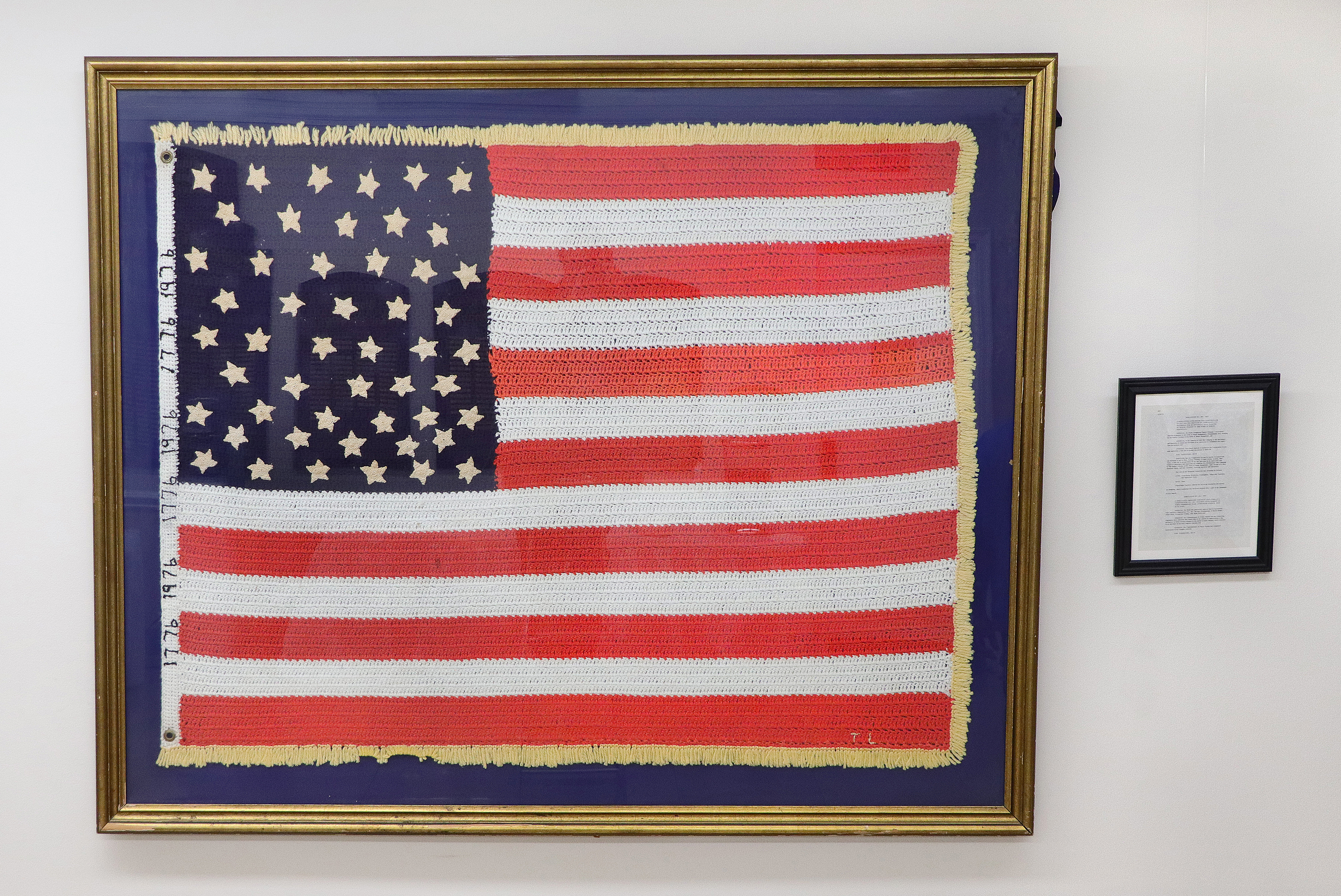 The crocheted American flag, originally created for the U.S. Bicentennial in 1976, displayed in its gold frame alongside a framed historical document.